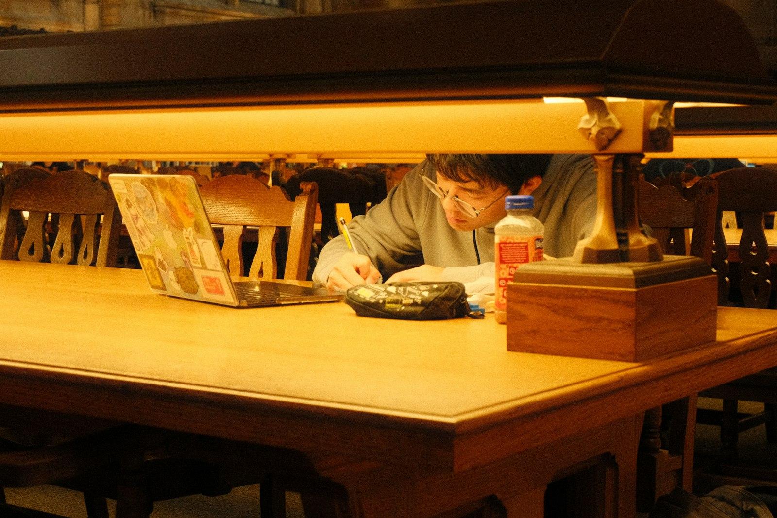 A focused student writing beside a laptop in a university library